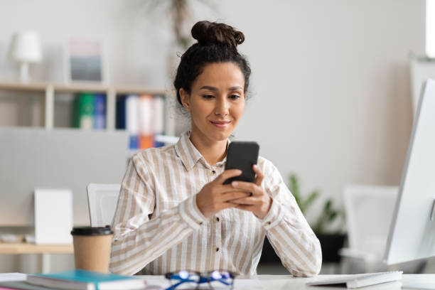 istock Smiling young female office worker using smartphone and reading message, sitting at table with pc in office interior 1478505671