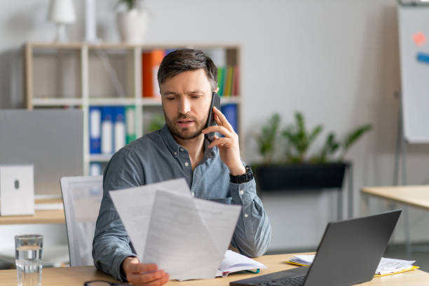 istock Handsome mature male manager sitting at office, reading documents and having phone conversation, copy space 1478506114