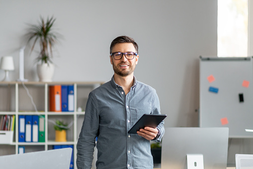 Portrait of happy mature office worker holding digital tablet, looking and smiling at camera, standing near his workplace. Successful male entrepreneur with modern device