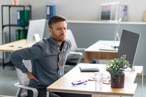 Exhausted mature businessman having acute back pain, massaging aching muscles while sitting at desk in office