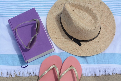 Beach towel with book, straw hat, sunglasses and flip flops on sand, flat lay
