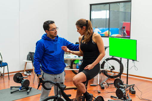 Latino man of average age of 35 dressed comfortably is inside his physical rehabilitation office for cyclists who does it through the technology of his laptop and technological devices, he attends to one of his clients, a Latina woman who will put her rehabilitation in her hands