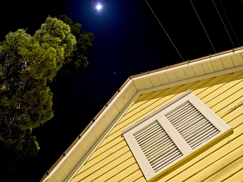 Moonlit night, a yellow house and shutter contrasts with the deep blue sky of moonlight and stars.