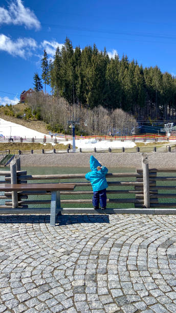 small two-year-old swings to throw a stone into a clear mountain lake small two-year-old child, boy, Toddler in a blue jacket with a sharp hood, cold early spring, walk, swings to throw a stone into a clear mountain lake, a wooden fence, snow on the slopes, a sunny day bukovel ski resort ukraine stock pictures, royalty-free photos & images