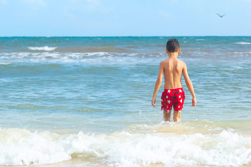 A boy in red swimming shorts are walking into the water in the ocean beach. Blue sky and waves on the background.