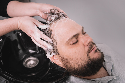 A young bearded man of Caucasian appearance at a reception at a barber shop. The process of cutting hair. Hairstylist washing client's hair in barber shop
