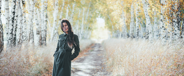 Sad beautiful girl with long natural black hair on bokeh background of autumn yellow leaves. Depressed girl in fall forest. Autumn apathy. Female emotional gloomy portrait in full growth. Faded tones.