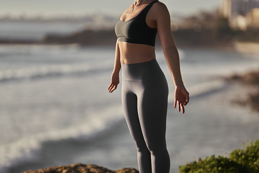 Crop unrecognizable fit woman in top and leggings standing on rocky coast in Tadasana pose with mudra hands while practicing yoga near wavy ocean at sunset