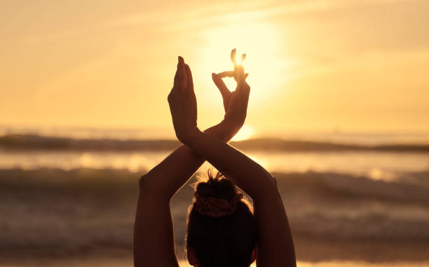 Back view of unrecognizable female with dark hair standing in front of powerful wavy ocean with raised Gyan mudra hands while practicing yoga against sunset sky