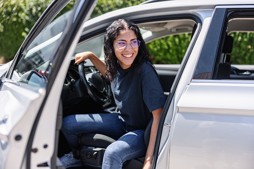 Happy young Latin female with glasses is stepping out of her parked car. The car door is open.