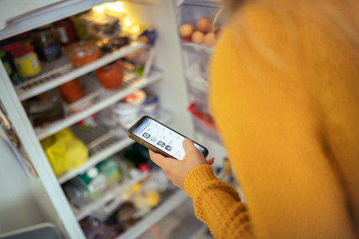 An unrecognizable woman opens the refrigerator while buying groceries via a mobile app and checking what else she needs to order
