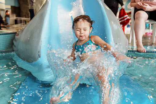 A cute preschool age multiracial girl of Pacific Islander descent closes her eyes as she splashes into the pool when riding down a water slide at a public pool.