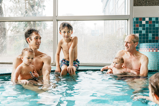 An active senior man playfully interacts with his multiracial grandchildren and adult son while spending a fun and relaxing day swimming at an indoor pool.