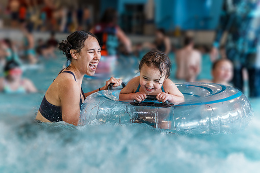 An Eurasian woman and her preschool age daughter laugh together while using an inflatable float ring in an indoor wave pool.