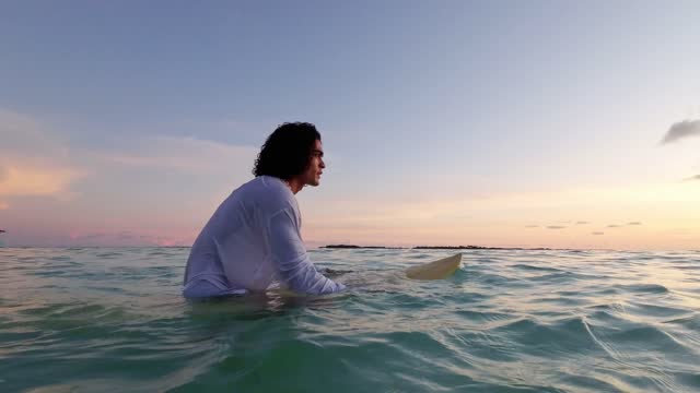 Young surfer sitting on the surfboard in ocean at the sunset