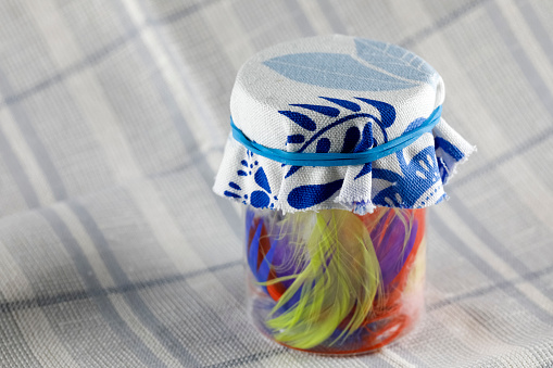 A small jar with colorful feathers inside is topped with a piece of fabric and secured with a blue rubber band. In the jar, among the feathers, there are little surprises, this is a surprise gift.