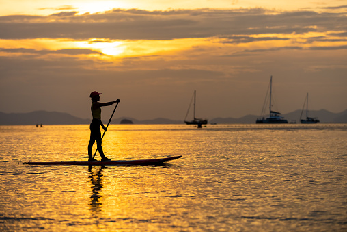 Asian mature woman standing on paddle board and rowing in the ocean at tropical island at sunset. Wellness woman enjoy outdoor lifestyle and water sports surfing and paddle boarding on summer vacation