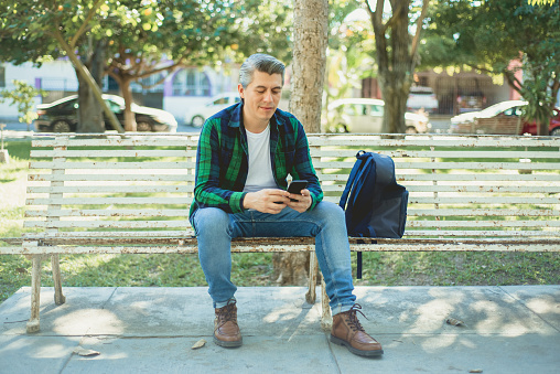 Gray-haired adult male in a park sitting on a bench looking at a cell phone and smiling.