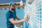 Bearded man spreads the cement grout on the pool tile to waterproof it, preparing pool for summer