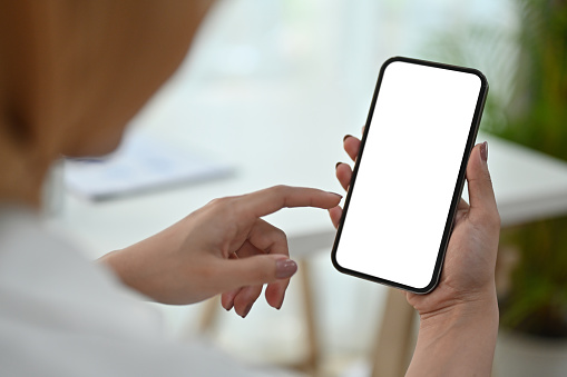 View over shoulder of woman hand holding smart phone. White empty screen for app or web site design.