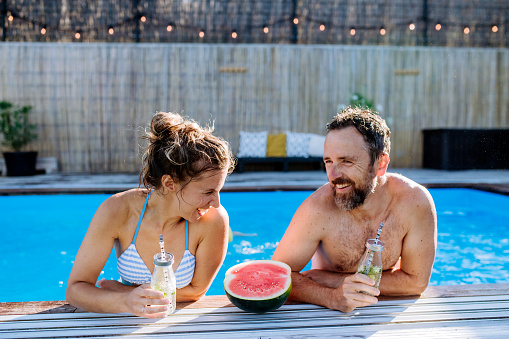 Happy couple enjoying summer time in a swimming pool, having drinks and watermelon.
