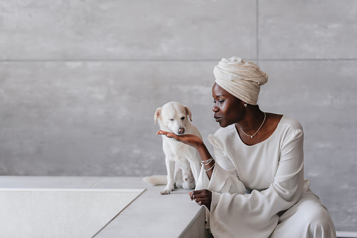 African woman in turban and white dress sitting at bathroom tenderly touching her white dog eyes closed. Adorable African American female home spending time with beloved pet. Pets and owners. Mockup.