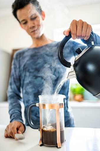 Mid adult person pouring hot water from kettle into french press coffee maker on kitchen counter at home
