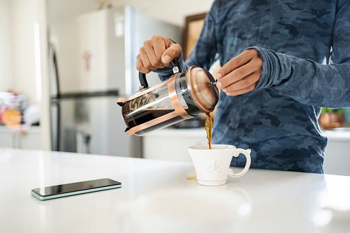 Cropped shot of unrecognizable person pouring coffee from french press into a cup on kitchen counter at home