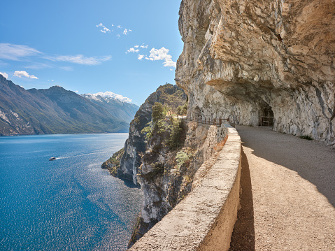 Wide-angle view of a scenic section of Strada del Ponale (Ponale Road), a former highway open to vehicles connecting Lake Ledro and the town of Riva del Garda, nowadays a panoramic route reserved to hikers and cyclists. The warm, bright light of a spring day, picturesque clouds, high, looming walls of rock, a narrow trail cutting into the side of the mountain, a ferryboat crossing the deep blue waters of the lake. The cliffs and slopes of Monte Baldo, and a long stretch of the eastern shore of Lake Garda can be seen on the background. High level of detail, natural rendition, realistic feel. Developed from RAW.