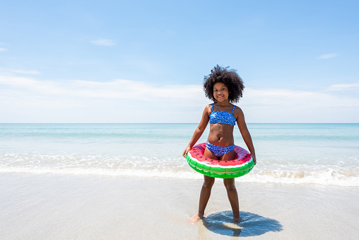 Little African girl in swimsuit with swim ring running and playing on tropical beach sea in sunny day. Child girl kid enjoy and fun outdoor lifestyle activity travel ocean on summer holiday vacation.