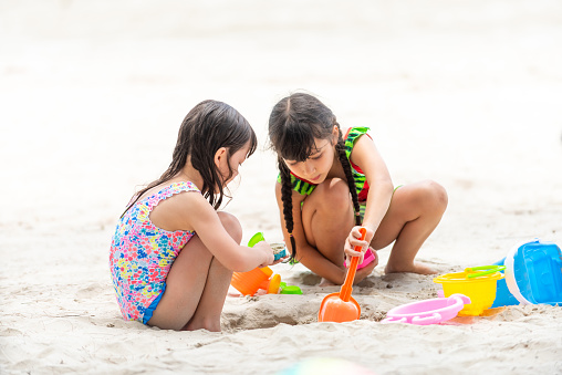 Little Asian child girl friends in swimsuit playing beach toy and build a sand castle together at tropical beach. Children kids enjoy and fun outdoor activity lifestyle travel ocean on summer vacation.