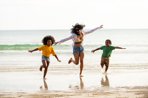 Happy African family mother and little daughter and son running and playing sea water together on the beach at summer sunset. Mom with child boy and girl enjoy outdoor lifestyle vacation at the sea
