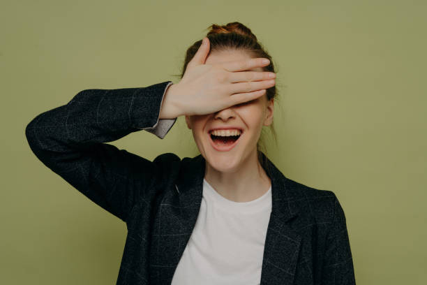 Laughing young female in casual clothes covering eyes with hand while waiting for surprise Laughing young female in casual clothes covering eyes with hand while waiting for surprise, smiling teenage girl keeping mouth opened with embarrassed expression, studio shot hair covering eyes stock pictures, royalty-free photos & images