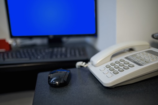 Desk with a black mouse and white telephone with a monitor screen on blue on the background and a keyboard also. Shallow depth of field