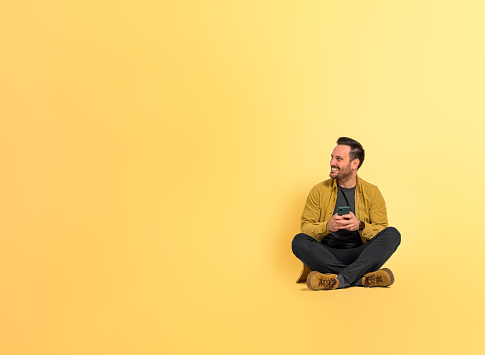 Full length of cheerful good looking mid adult man text messaging over smart phone and looking away while sitting with legs crossed isolated on yellow background