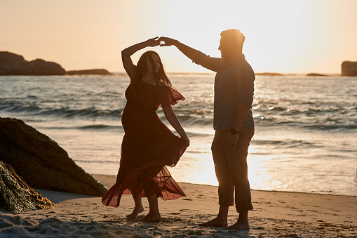 Couple, pregnancy and dance in sunset at beach with silhouette, peace and relax with sound of waves. Man, woman and holding hands with dancing, support and sunshine for holiday, wellness or freedom