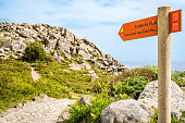 A signpost directs hikers to PR 3 Trilho da Fóia and PR 5 Percurso das Cascatas trails, leading to the sunlit Fóia mountain top with the distant haze-wrapped city of Portimão visible in the background
