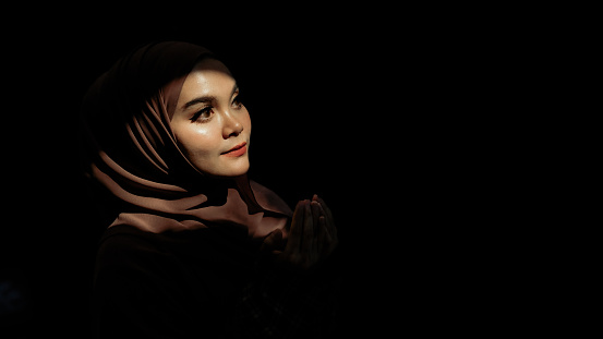 Portrait of a Muslim woman in hijab praying on dark background. Space for text