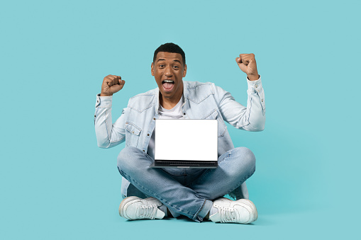 Cheerful excited young african american guy with open mouth shout, make victory gesture, show laptop with empty screen isolated on blue background. Gadget for work, study, win and success gesture