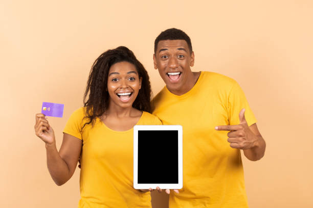 istock Happy black man pointing at empty tablet screen, his girlfriend holding credit card on peach studio background, mockup 1478730403