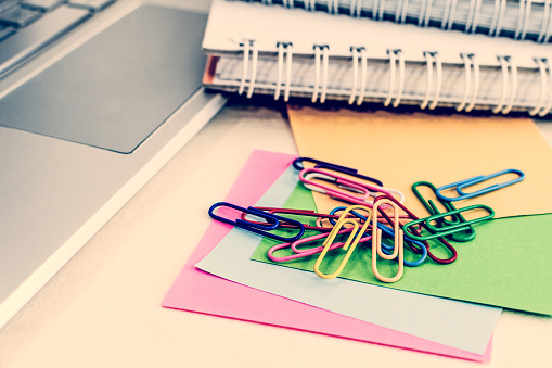 A closeup view of notebooks, colorful sticky notes, and paper clips, next to a laptop, on a desk