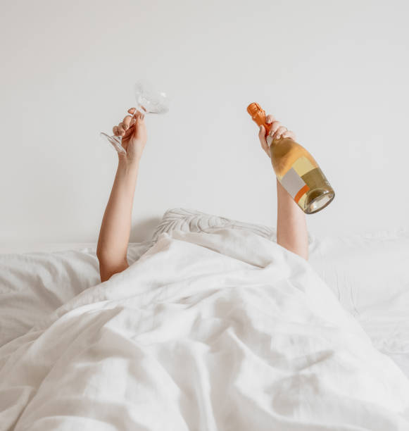 ベッドの白いシーツの下に横たわりながらグラスとシャンパンのボトルを持つ人 - champagne flute champagne glass alcohol ストックフォトと画像