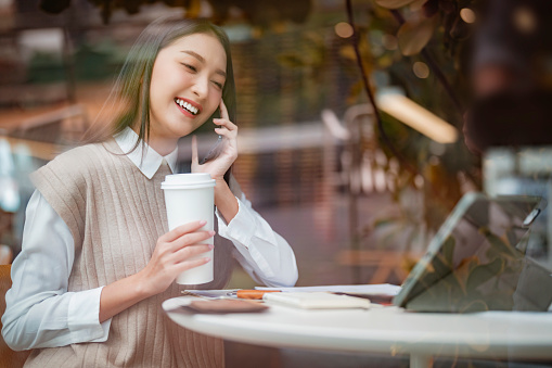 casual smart asian female entrepreneur small business working at coworking space hand using smartphone and tablet business talking consulting shooting through window glasses garden reflecting view