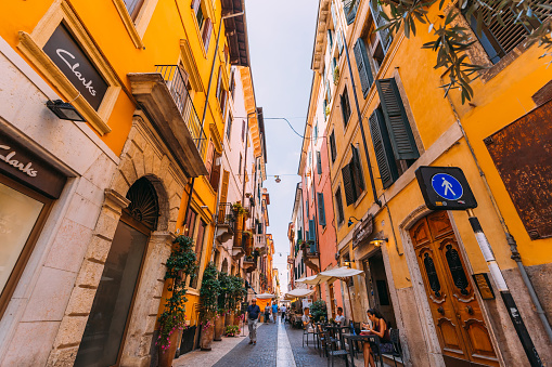 Verona, Italy - July 4, 2018 cozy narrow street between two buildings. cafe with bar parasols. branches of a tree with green leaves on the foreground