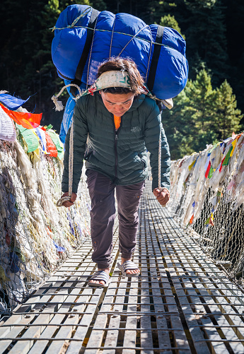 Sherpa porter carrying  expedition kit bags and equipment using a traditional head band across a wire bridge deep in the mountain wilderness of the Sagarmatha National Park, Himalayas, Nepal.