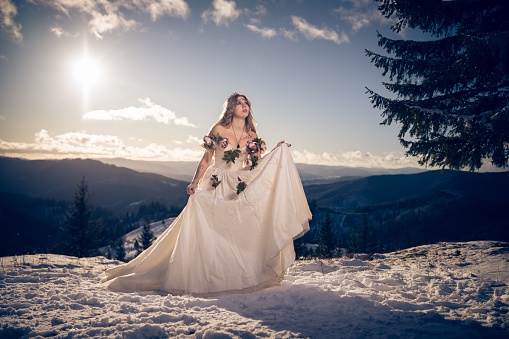 A beautiful young female in a mystical wedding dress with pink flowers posing on a snowy mountain