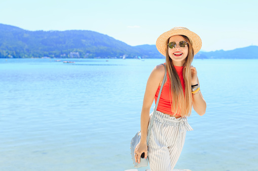 The beautiful girl in stylish clothes and sunglasses poses. happy and smiles. water of the lake and slopes of mountains on a background