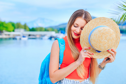A stylish girl with a backpack holds the boater in hands and closes the half face. lake and mountains