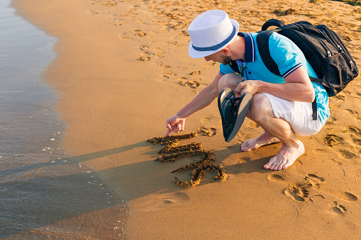 guy in summer clothes and a backpack writes the word on wet sand. sandy beach and the sea