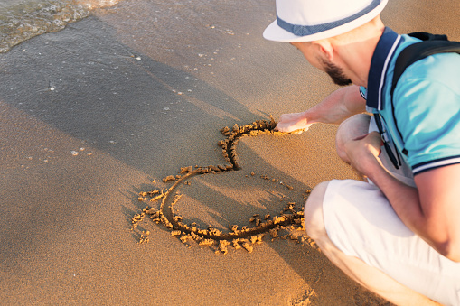 guy in a boater draws a heart symbol on wet sand near the sea